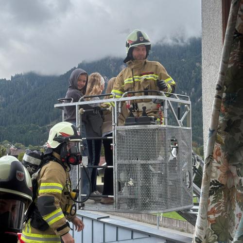 Im VOrdergrund steht ein Feuerwehrmann mit Atemschutzmaske am Flachdach des hinteren EInganges, dahinter der Bergekorb der Drehleiter in dem sich 2 Kinder und ein Feuerwehrmann befinden