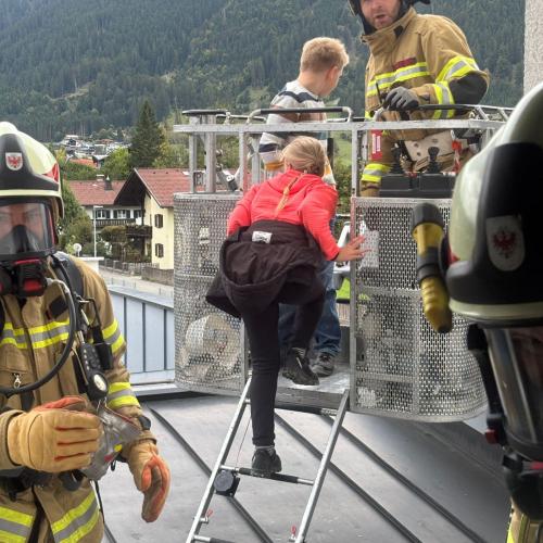 Am Flachdach des hinteren Einganges der Schule steht der Bergekorb der Drehleiter und zwei Feuerwehrmänner helfen einem Kind beim "einsteigen"
