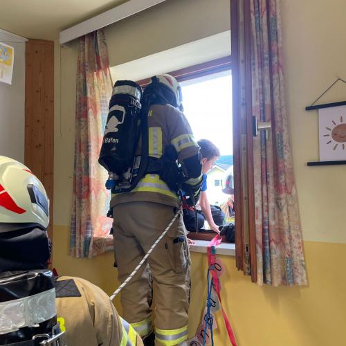 EInFeuerwehrmann steht beim offenen Fenster (1.Stock) und hilft einem Kind die Leiter hinuter, ein zweiter sichert mit einem Seil