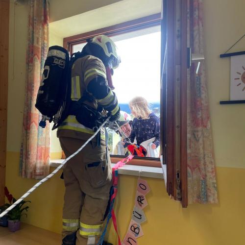 EIn Feuerwehrmann mit Atemschutzmaske steht im Klassenzimmer beim offenen Fenster und hilft einem Kind auf die Leiter