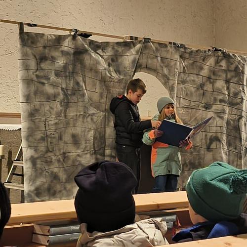 2 Kinder lesen aus einem großen Buch in der Kirche vor einer Kulisse, die ein Tor darstellt einen Teil der Martinslegende vor