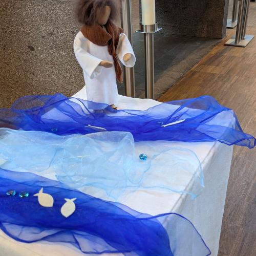 Eine Figur - Puppe - auf einem Tisch , umgeben von blauern Tüchern auf denen kleine Fische liegen