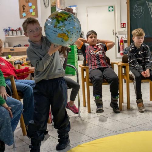 Ein Kind bewegt sich mit einem Globus in der Hand um ein großes, rundes, gelbes Tuch das am Boden liegt und die Sonne darstellen soll