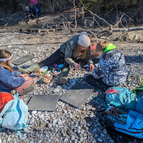 3 Kinder sitzen auf der Schotterbank, auf Sitzunterlagen und basteln mit Sabine von Natopiaein Bibermobile