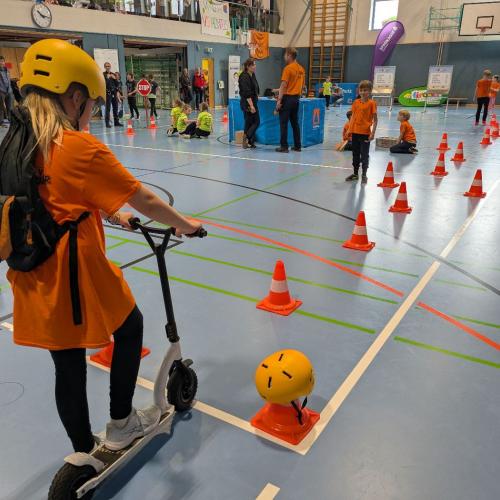 Ein Kind mit orangem T-Shirt und gelbem Helm startet einen Geschicklichkeitsparcour mit einem Scooter in der Turnhalle
