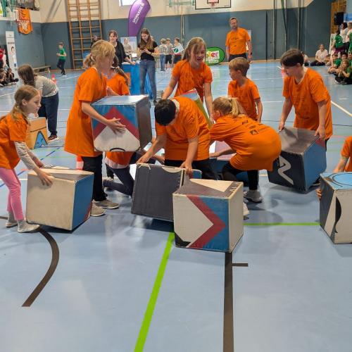 Die Kinder der 3a in orangen T-Shirts bauen aus Schaumstoffwürfeln ein Gefahrenstoffsymbol zusammen