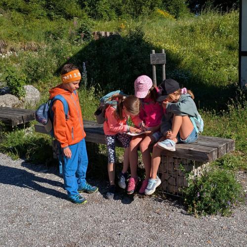 3 Kinder sitzen auf einer Bank, ein Bub steht daneben, sie lesen im Heft den Text für das Rätsel.