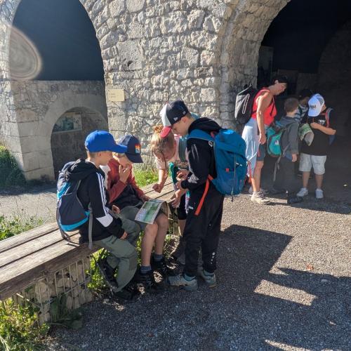 Eine andere Gruppe Kinder sitzt auf einer Bank und liest das Rätsel im Heft, im Hintergrund steht die 3. Gruppe.