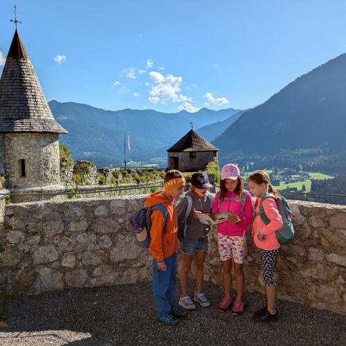 4 Kinder stehen bei einer Mauer, links befindet sich ein kleines Türmchen im Hintergrund Berge und strahlend blauer Himmel