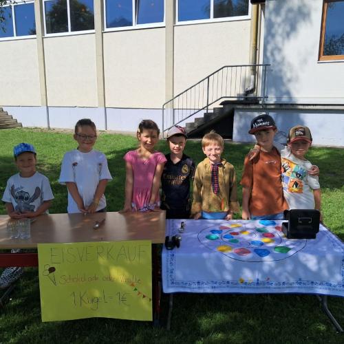 7  Kinder der 1 Klasse stehen hinter dem vorbereiteten Tisch zum Eisverkauf im Schulhof im Schatten der Birke