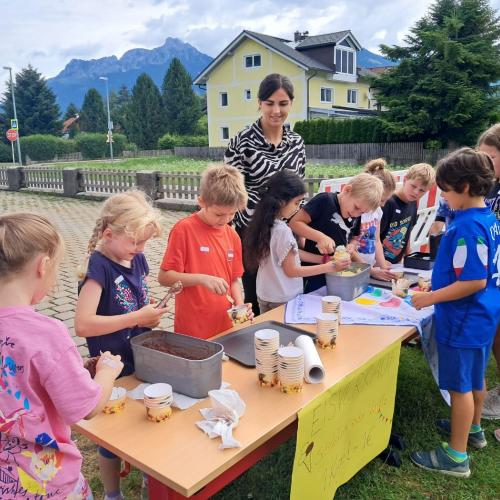 7 Kinder der 1. Klasse stehen hinter dem Verkaufstisch und kassieren das Geld, verteilen das EIs in Becher und geben Löffel dazu, 3 Kinder stehen an, um ein Eis zu kaufen, hinter den Kindern steht die Klassenlehrerin der 1. Klasse.