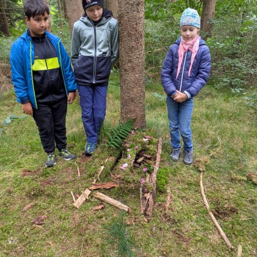 3 Kinder stehen stolz vor ihrem aus Moos und Zweigen gebautem Mäusehäuschen