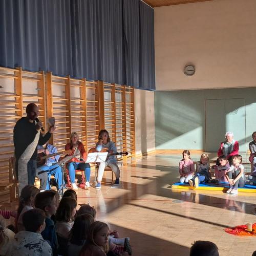 Die Lehrerinnen, die mit der Gitarre begleiten sitzen am Rand des Turnsaals, neben ihnen steht der Diakon Patrick Gläffe
