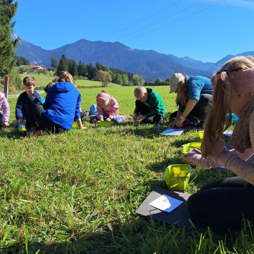 Kinder sitzen auf einer Wiese am Boden - strahlend blauer Himmel über einer Bergkette im Hintergrund