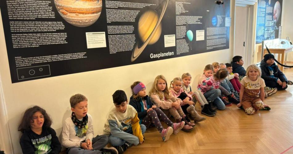 Schulkinder sitzen am Boden vor einer großen Tafel über die Planeten