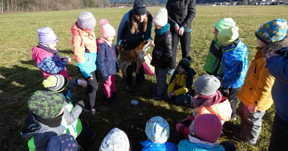 Auf einer Wiese stehen eine Gruppe Kinder im Kreis um die 2 Naturpädagogen von Natopia