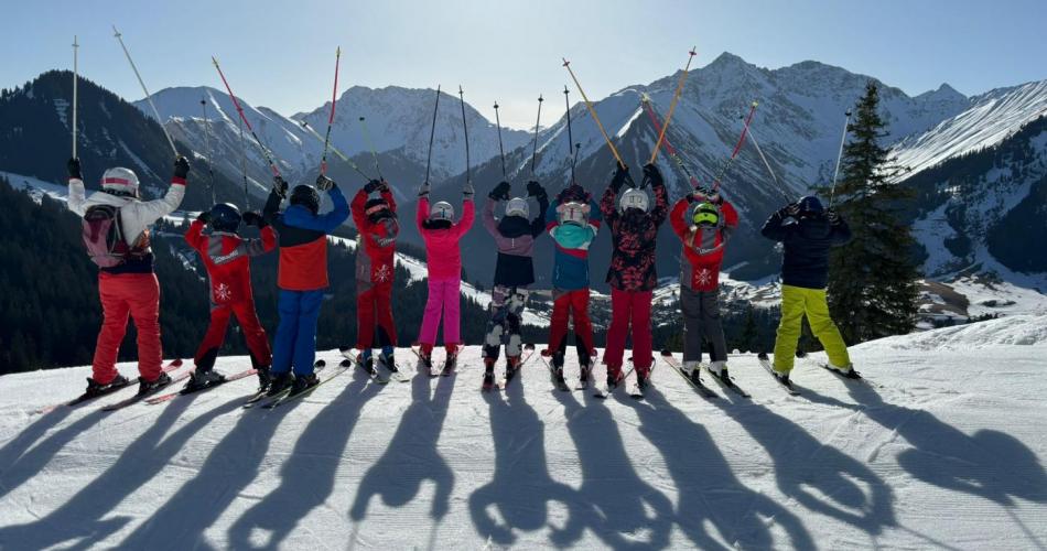 9 Kinder stehen mit ihrer Lehrerin in bunten Schanzügen bei strahlend blauem Himmel vor der Bergkulisse in Berwang