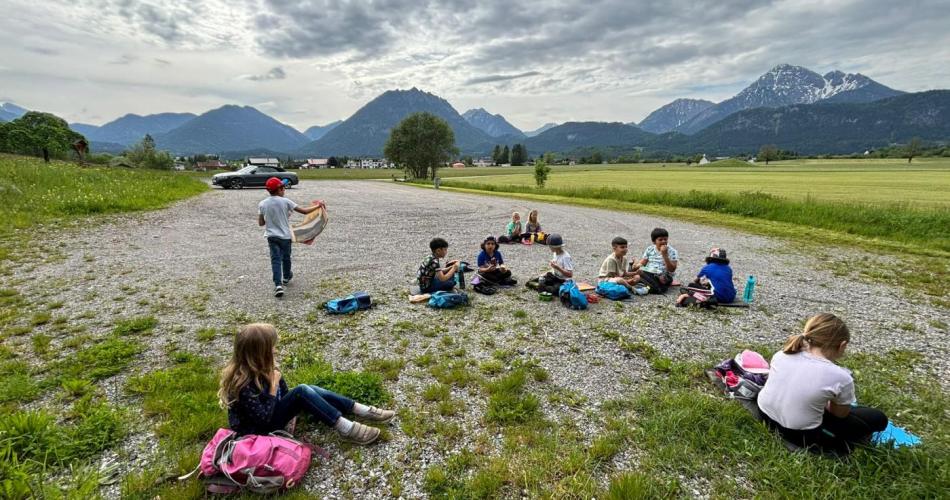 Kinder sitzen zum Jausnen auf einem Schotterplatz im Hintergrund die Berge des Talkessels Reutte