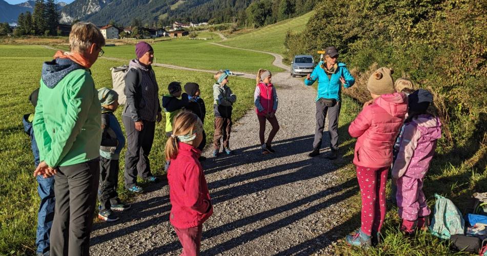 Auf einem Schotterweg am Waldrand stehen alle Kinder der Klasse im Kreis; strahlend blauer Himmel, im Hintergrund Berge