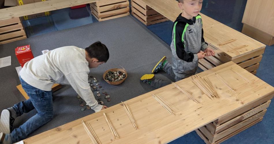 Ein Kind hat auf der Holzbank in der Klasse 100 Schaschlikstäbchen gezählt und gebündelt, ein anderes Kind legt 100 mit Steinen am Teppich.