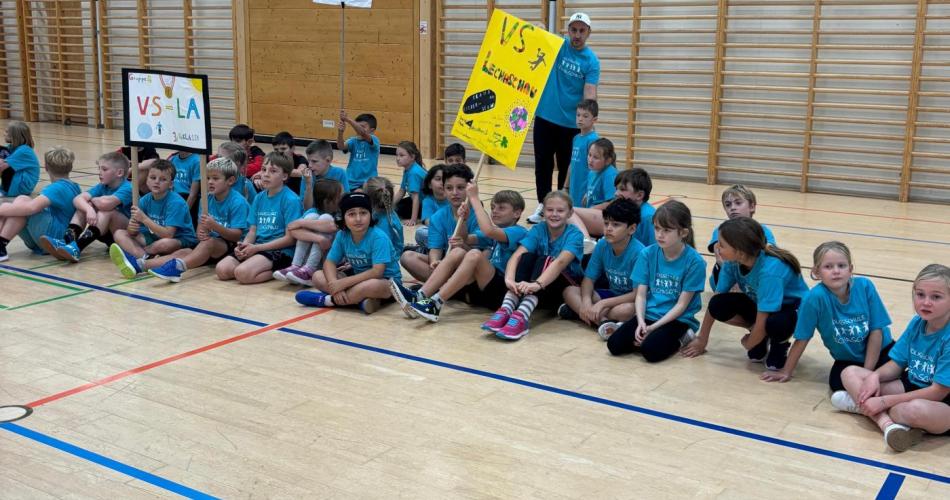 ca. 40 Kinder der Volksschule Lechaschau sitzen mit blauen Schul T-Shirts am Boden der Turnhalle Reutte im Hintergrund die Sprossenwände