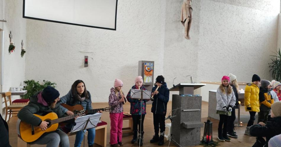 Kirche Lechaschau. im ALtarraum sitzen 2 Lehrerinnen mit Gitarren, daneben stehen drei Kinder mit Blockflöten, danach stehen noch ein paar Kinder , die etwas vorgelesen haben.