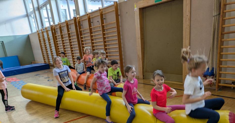 Die Mädhen der Klasse sitzen im Turnsaal auf einer gelben Gummihüpfwurst