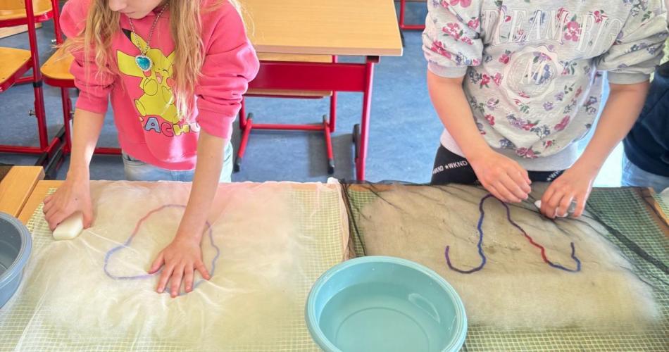 2 Kinder stehen bei einem Schultisch - sie haben eine Wasserschüssel vor sich, Folie auf einem Handtuch und filzen mit Wolle