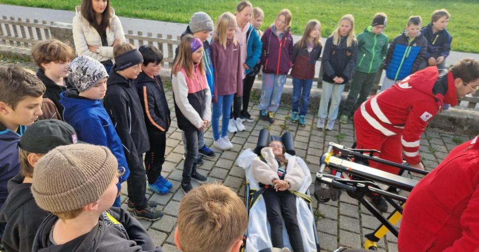 SchülerInnen stehen mit der Klassenlehrerin im Schulhof, ein Kind liegt auf der Rettungsliege - die Rettung ist zu Besuch und stellt das AUto und Geräte vor