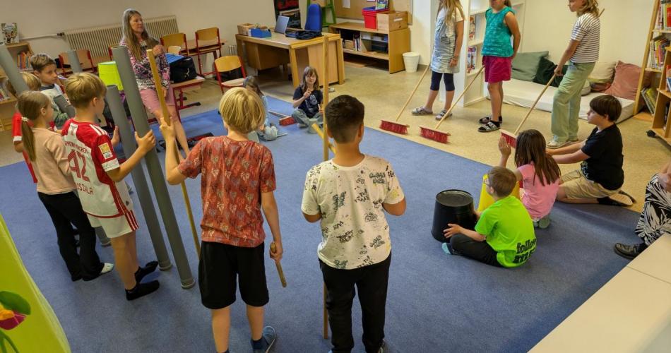 Die Kinder der Klasse sind in der Bibliothek, stehen im Kreis mit ungewöhnlichen Rhythmusinstrumenten, wie Besen, Eimern, Holzstäben, Rohren..