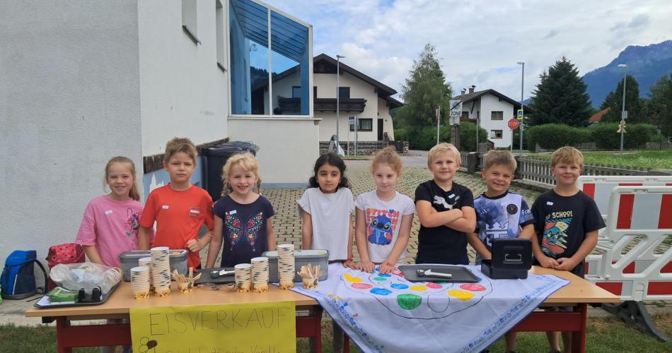 8 Kinder der 1. Klasse stehen hinter dem dekoriertem vorbereitetem Tisch für den Eisverkauf im Pausenhof